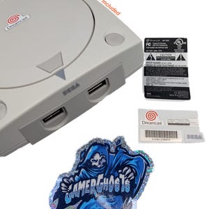 May include: A grey Sega Dreamcast console with a sticker that says "Dreamcast" and a sticker that says "Gamer Ghosts". Also included are two other stickers with text and logos. The text "DreamCase Not Included" is also present.
