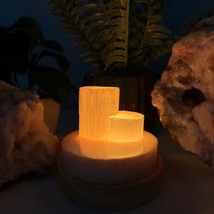 May include: A cylindrical selenite lamp featuring two vertical pillars of varying heights that emit a warm, glowing orange light. The translucent, fibrous crystal structure is mounted on a circular base, creating a soft, ambient illumination.