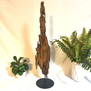 May include: A tall, weathered piece of driftwood, mounted on a black metal stand. The wood is a mix of brown and tan hues, with a rough, textured surface. The sculpture is a natural, organic home decor accent.