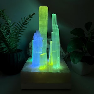 May include: A vertical selenite crystal lamp featuring multiple translucent, pillar-like formations mounted on a rectangular base. The crystals are illuminated from within, emitting vibrant neon green and blue light glows throughout the structure.