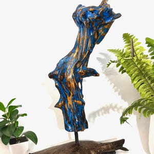 May include: A blue and gold abstract wooden sculpture. The artwork features a vertical, textured form with a vibrant blue and gold color scheme. The sculpture is mounted on a dark wooden base and is displayed against a white background, with green plants in the background.