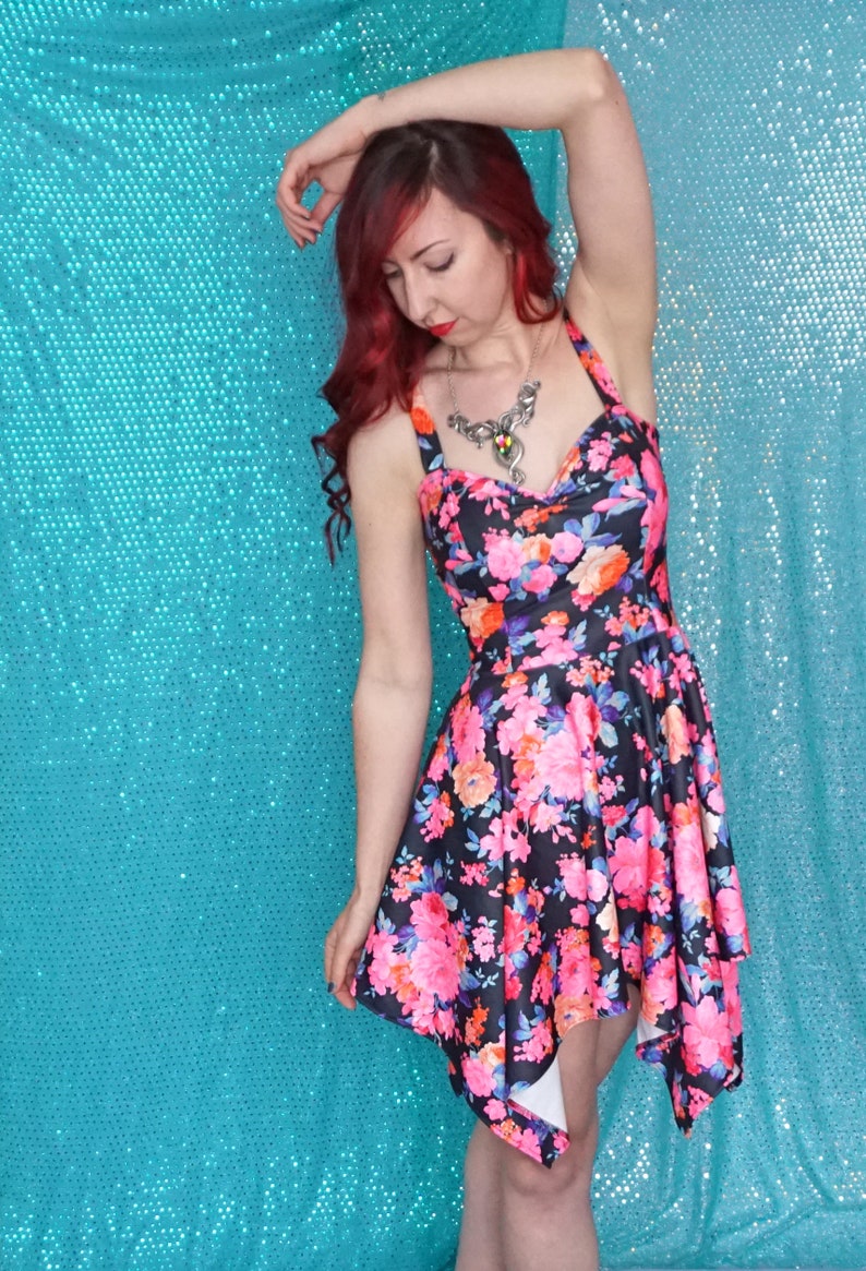 May include: A woman wearing a black and pink floral print dress with a silver necklace. The dress has a high neckline and a short, asymmetrical hemline. The woman is standing in front of a turquoise background.