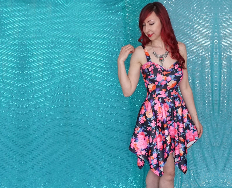 May include: A woman wearing a black and pink floral print dress with a sweetheart neckline and a short, asymmetrical hemline. She is standing in front of a turquoise sparkly background.