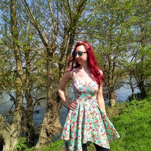 May include: A woman with red hair wearing a floral print dress and sunglasses stands in front of a river and trees. She is wearing a floral headband.