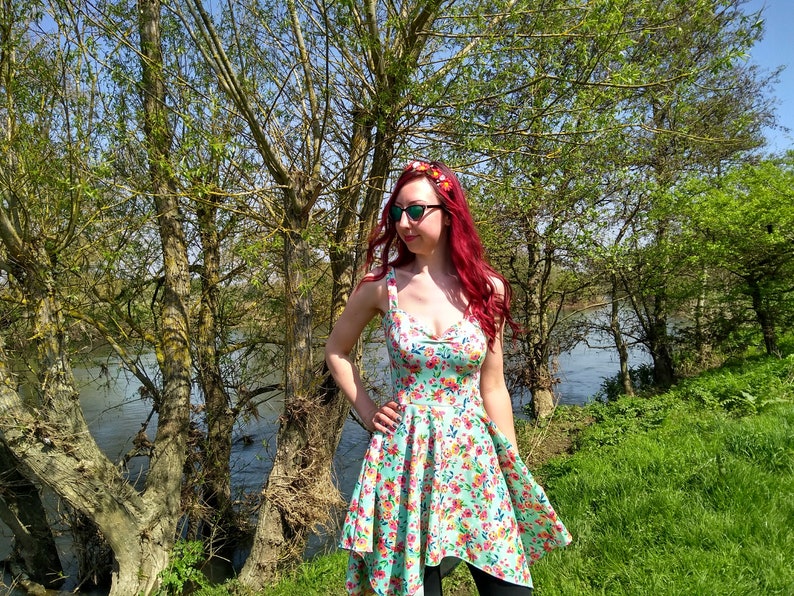 May include: A woman with red hair wearing a floral print dress and sunglasses stands in front of a river and trees. She is wearing a floral headband.