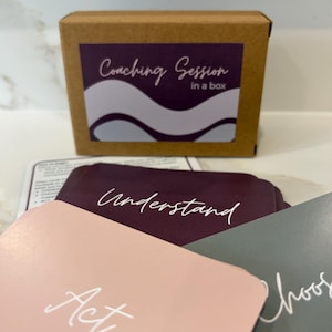 May include: A brown cardboard box with a purple label that reads "Coaching Session in a box." Several cards are visible, with the words "Act," "Understand," and "Choose" printed on them in white script.