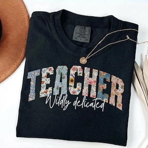 Puede incluir: Una camiseta negra de manga corta con la palabra TEACHER en un patrón floral con varias flores y colores. Debajo de la palabra, el texto Wildly dedicated está impreso en letra cursiva blanca. La camiseta tiene una etiqueta Comfort Colors.