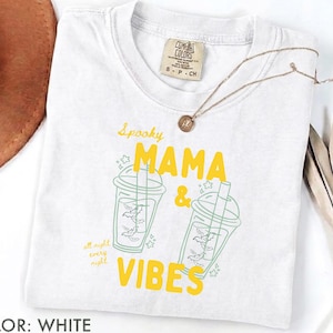 Puede incluir: Una camiseta blanca con un estampado gráfico de dos vasos de bebida helada con murciélagos dentro. El texto dice Spooky MAMA & VIBES y all night every night en amarillo y verde. La camiseta tiene una etiqueta de la marca Comfort Colors.