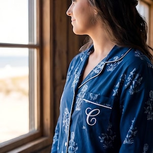 Könnte beinhalten: Marineblaues Pyjam Oberteil mit weißem Blumenmuster und weißer Paspelierung. Das Oberteil hat eine Brusttasche mit einem weißen, gestickten Initialen. Die Person schaut aus einem Fenster auf den Strand.