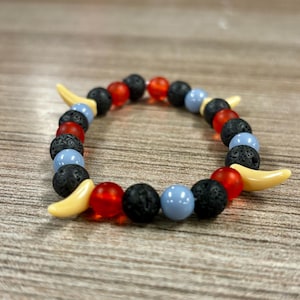 Pode incluir: Uma pulseira de contas com uma variedade de contas coloridas. A pulseira inclui contas de lava pretas, contas redondas vermelhas e azul claro e contas em forma de banana. A pulseira é exibida em uma superfície de madeira.