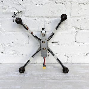 May include: A quadcopter drone frame featuring a central metallic chassis with four extended arms. Each arm terminates in a black motor. A small camera is mounted on the front, and a yellow electrical connector hangs from the rear of the frame.