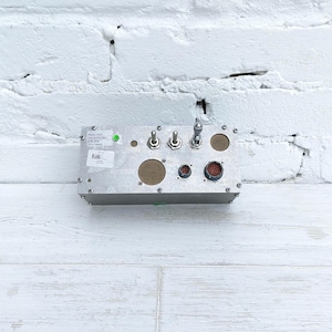 May include: A rectangular, silver-colored electronic control box with various switches, dials, and connectors. The box has a series of toggle switches, circular buttons, and a multi-pin connector. The surface has a few labels and a green indicator light.