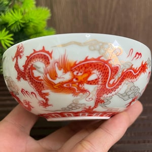 Könnte beinhalten: Eine weiße Keramikschale mit einem leuchtend roten und orangefarbenen Drachendesign. Der Drache ist aufwendig detailliert, mit goldenen Akzenten und Wolkenmotiven. Der Rand der Schale hat einen goldenen Besatz.