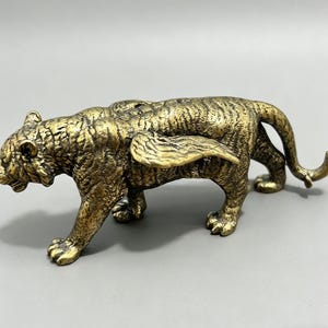 Può includere: Una statuetta di tigre alata color bronzo, in piedi sulle quattro zampe. La tigre ha una texture di pelliccia dettagliata ed è in posa mentre cammina. Le ali sono dettagliate e aggiungono un elemento unico alla scultura.