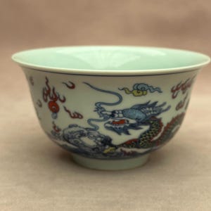 May include: A white ceramic bowl featuring a blue dragon motif. The dragon is detailed with blue, red, and green colours. The bowl has a rounded form, a gently flared rim, and a light blue interior.