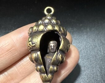 Op de afbeelding: Een bronzen hanger met een Boeddha-beeldje in een schelpvormige behuizing. De schelp heeft ingewikkelde details en een lus om op te hangen. De Boeddha zit, en de hanger heeft een verouderd uiterlijk.