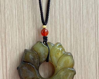 Op de afbeelding: Een cirkelvormige, olijfgroene stenen hanger uitgesneden in de vorm van een bloem met meerdere bloemblaadjes. De hanger hangt aan een zwart gevlochten koord, geaccentueerd door een kleine goudkleurige kraal en een rode, doorschijnende kraal.