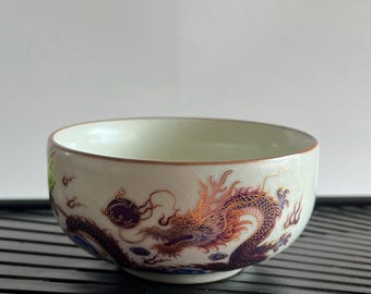 May include: A white ceramic bowl with a detailed dragon design in shades of purple, orange, and gold. The bowl has a light green interior and a brown rim. The dragon is depicted with intricate scales and a dynamic pose.
