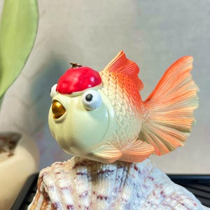 May include: A whimsical, hand-painted goldfish figurine with a red cap, perched atop a seashell. The fish features orange fins and tail, with a cream-colored body and large, expressive eyes. A unique decorative item.