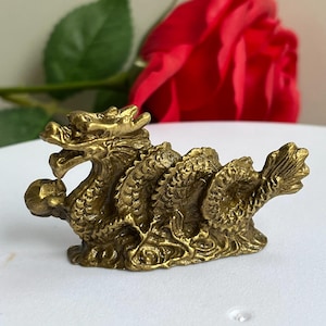 May include: A small, metallic gold-coloured dragon figurine featuring intricate scale detailing along its coiled body. The dragon is depicted in a horizontal, serpentine pose with its head turned slightly, holding a small spherical object in its mouth.
