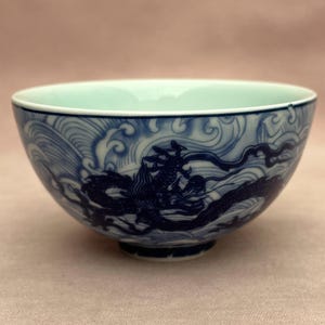 Könnte beinhalten: Eine blau-weiße Keramikschale mit einem Drachenmuster. Die Schale zeigt eine detaillierte Illustration eines Drachen inmitten von wirbelnden Wellen. Das Innere der Schale ist hellgrün und bildet einen Kontrast zum blau-weißen Äußeren.