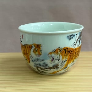 Könnte beinhalten: Weiße Keramikschale mit einer detaillierten Illustration von zwei Tigern. Die Tiger sind orange und schwarz, vor dem Hintergrund einer verschneiten Landschaft und Bäumen. Die Schale hat eine glatte, glänzende Oberfläche.