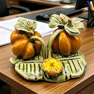 Pode incluir: Uma peça decorativa de cerâmica com duas abóboras laranja brilhantes com caules e folhas verdes, sobre uma base grande de folha verde texturizada. Uma abóbora menor com listras amarelas e laranja está situada no centro da base.