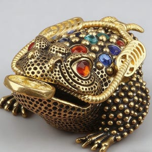 May include: A detailed, gold-colored frog figurine with a coin in its mouth. The frog's body is adorned with small, round details and colorful gemstones. The figurine has a textured surface and a decorative coin design.
