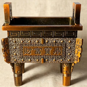 May include: A rectangular bronze-colored metal incense burner featuring four legs, two upright handles, and a textured surface with a geometric border. The front panel displays the Chinese characters 財富寶鼎 in relief, surrounded by a decorative pattern.