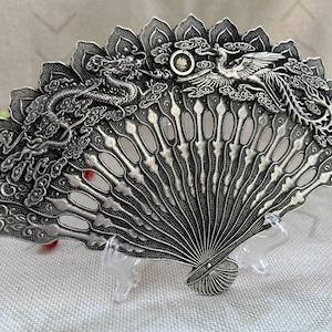 May include: A silver-toned metal hand fan featuring intricate embossed designs of a dragon and a phoenix amidst clouds. The fan has a scalloped top edge and a series of vertical, open-work slats radiating from a central pivot point at the base.