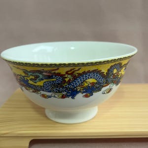 Könnte beinhalten: Weißer Keramiknapf mit Sockel, mit einem gelb-blauen Drachendesign. Die Schüssel hat einen dekorativen Rand und steht auf einer Holzoberfläche. Das Drachendesign enthält rote und blaue Akzente.