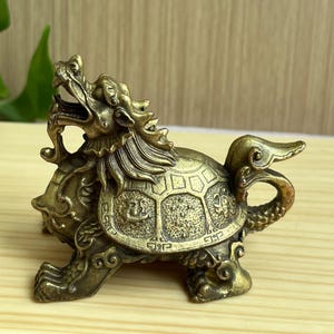 Puede incluir: Una figura de metal color latón de una criatura mítica con cabeza de dragón y cuerpo de tortuga. La criatura presenta una boca abierta, un caparazón texturizado con patrones grabados y una cola curva, apoyada sobre cuatro patas.