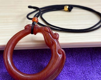 Op de afbeelding: Een roodbruine hanger van gesneden steen in de vorm van een cirkelvormige draak, hangend aan een zwart koord. De hanger is voorzien van een kleine oranje kraal boven de drakenfiguur en een goudkleurige kraal op het koord voor een samenhangend geheel.