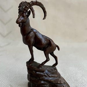 Puede incluir: Una escultura de color bronce de una cabra montesa con grandes cuernos curvados. La cabra está de pie sobre un afloramiento rocoso, su cuerpo ligeramente inclinado. La escultura tiene detalles y un acabado liso.