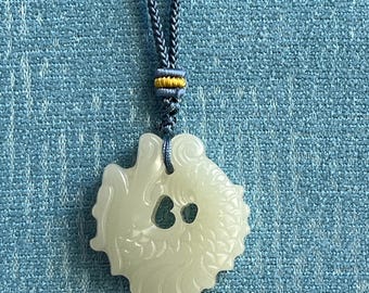 Op de afbeelding: Een lichtgroene, doorschijnende stenen hanger uitgesneden in een cirkelvormige drakenfiguur met gedetailleerde schubben en een centrale uitsparing. De hanger hangt aan een blauw gevlochten koord met een kleine, decoratieve geel-blauwe schuifknoop.