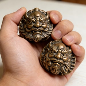 May include: Two bronze-colored, round decorative objects, each featuring a detailed, stylized face with horns, a beard, and an intense expression. The objects are held in a hand, showcasing their intricate design and metallic finish.