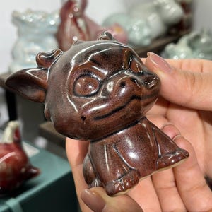 Pode incluir: Uma estatueta de cerâmica marrom de um dragão sorridente com olhos grandes, chifres pequenos e orelhas pontudas. O dragão está sentado, com as pernas dobradas. A estatueta tem um acabamento brilhante e é segurada em uma mão.