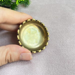 以下が含まれることがあります： 真鍮色の小さな円形の金属製皿で、縁はスカラップ状になっています。皿の内側は滑らかでわずかに光沢のある温かみのあるゴールドトーンで、外側の縁はより暗いアンティーク調の仕上げが施されています。