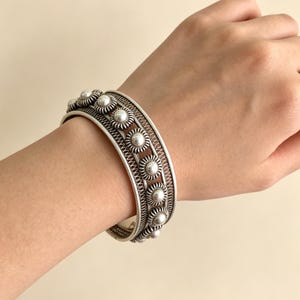May include: A silver cuff bracelet with an ornate design. The bracelet displays alternating rows of floral-style decorations and detailed openwork patterns. The floral elements have a central pearl-like bead. The bracelet is worn on a wrist.