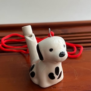 Puede incluir: Silbato de cerámica con forma de perro dálmata. El silbato blanco tiene manchas y orejas negras. Se adjunta un cordón rojo. El silbato mide unos 7 cm de largo y está sobre una superficie de madera.