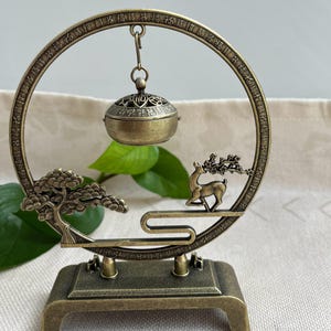 May include: An antique bronze-colored incense burner with a circular frame. The frame features Chinese characters and supports a hanging incense holder and a decorative deer and tree scene. The base is rectangular with two small posts.