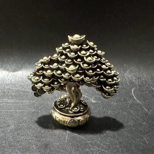 May include: A bronze-colored miniature tree sculpture. The tree's foliage is composed of numerous coin-shaped elements, and it is set on a round base. The sculpture has a detailed, textured appearance and is displayed against a dark background.