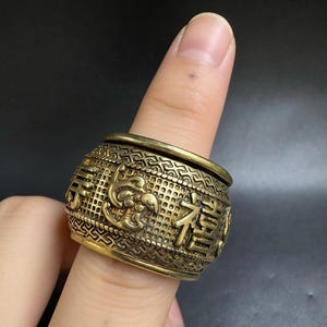 Può includere: Un anello in ottone in stile antico con intricati motivi in rilievo e caratteri cinesi. L'anello è cilindrico con un bordo superiore e inferiore leggermente rialzato. L'anello è indossato su un dito.