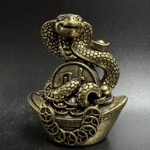 Peut inclure: Statue de cobra de couleur bronze avec un motif de pièce et de lingot. Le cobra est posé avec sa capuche évasée, au sommet d'une grande pièce. La base est un lingot en forme de bateau, avec des pièces décoratives sur les bords.
