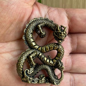 May include: A small, antique-style brass figurine of a coiled dragon with detailed scales, a horned head, and an open mouth. The dragon is depicted in a serpentine pose, clutching a small sphere with one claw, featuring a dark, weathered finish.