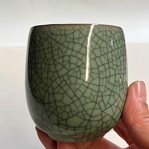 Puede incluir: Una taza de té de cerámica verde celadón con un patrón de esmalte craquelado. La taza tiene una forma redondeada y un borde marrón oscuro. La superficie está cubierta por una red de finas líneas oscuras.