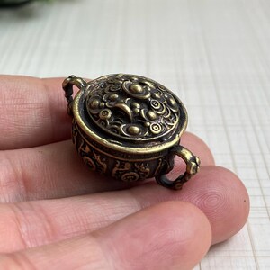 May include: A small, round, antique-style brass incense burner featuring intricate relief carvings on the lid and body. The vessel has two small, ornate side handles and a dark, weathered patina finish with polished highlights on the raised details.