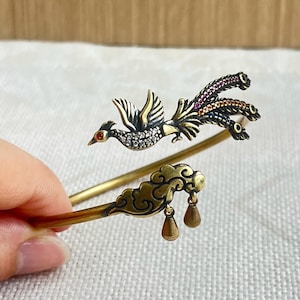 May include: A gold-toned metal bracelet featuring a detailed phoenix design. The phoenix has a red eye and is adorned with clear and colorful gemstones. The bracelet includes a cloud and teardrop charm.