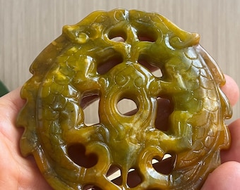 Op de afbeelding: Een cirkelvormige, doorschijnende geelgroene jade hanger met een ingewikkelde, opengewerkte gravure van een drakenmotief. Het ontwerp bevat een centrale cirkelvormige opening en gedetailleerde schubpatronen, met gladde, gepolijste randen.