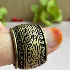 May include: A wide, antique brass ring with an intricate design. The ring features a repeating geometric pattern and a central motif of a stylized bird or mythical creature. The ring is approximately 1 inch wide.